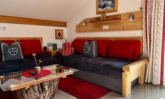 A cozy living room setup with a dark sofa and red cushions. The table is made of wood, and the walls are adorned with rustic decor.