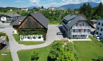 Two buildings in a green environment with a view of the mountains. The left house has a wooden facade with plants, while the right house has balconies.