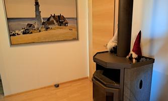 A cozy room with a modern stove and a large painting of a lighthouse. In the background, a wooden door is visible, and a small decorative figure stands on the stove.