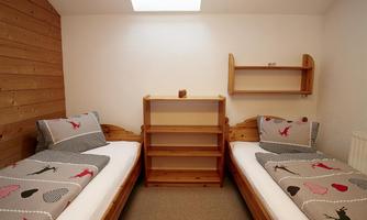 A simple bedroom with two single beds and wooden furniture. The walls are light and there is a shelf between the beds.