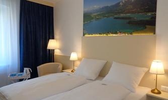 A cozy hotel room with two single beds and soft lighting. On the wall hangs a picture of a picturesque landscape with a lake and mountains.