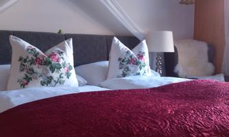 A cozy bed with flower-decorated pillows and a red blanket. In the background, there is a nightstand with a lamp.