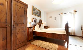 A cozy bedroom with a double bed and a wooden wardrobe. The walls are light and there are windows that let in plenty of light.