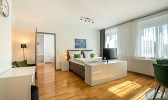A modern bedroom with a large bed, a television, and a desk. The room has large windows that let in plenty of daylight.