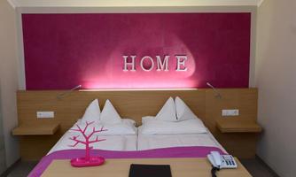 A modern bedroom with a large double bed and bright linens. A decorative element with the word "HOME" in striking letters hangs on the wall.