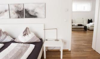 A modern bedroom with a double bed and a bedside table. In the background, the living room is visible, which is bright and inviting.