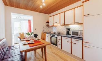 A modern kitchen with wooden ceilings, bright cabinets, and a dining table. The table is set, and there is a large window opening that lets in daylight.