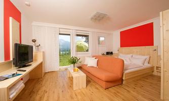 A modern room with an orange sofa and a wooden bed. Large windows offer a view of nature.