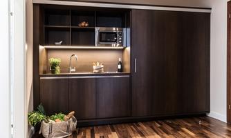 A modern kitchen unit with dark wooden cabinets and a lit workspace. On the counter, there are plants and a bottle of wine.