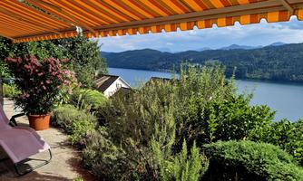 A beautiful view from a terrace over a lake, surrounded by green plants and mountains in the background. The awning is orange striped and provides pleasant shade.