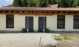 A modern house with two doors and a wooden overhang. In the background, there are trees and a hilly landscape.