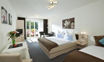 A modern hotel room with two beds and a cozy seating area. Large windows offer a view of a garden and bring natural light into the room.