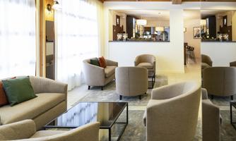 A modern living room with beige sofas and a glass table. Bright windows and an open bar in the background create an inviting atmosphere.