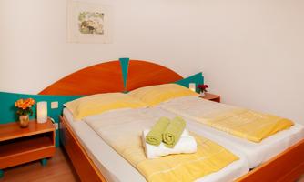 A cozy bedroom with a double bed and colorful bedding in yellow and white. Next to the bed are nightstands with flowers and towels.