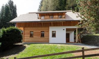 A modern wooden house with white walls and a sloped roof. It stands in a green environment with trees and a fence.