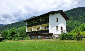 A large, two-story house with balconies, surrounded by green meadows and trees. The mountains in the background are partially wooded and the sky is cloudy.