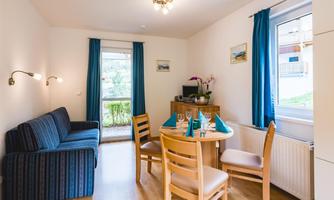 A cozy living space with a blue sofa and a dining table. Large windows provide bright light and a pleasant atmosphere.