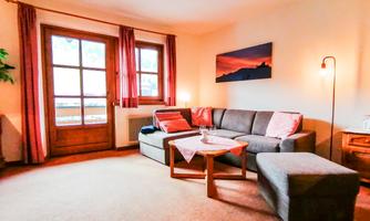 A cozy living room with a gray sofa and a coffee table. Large windows with red curtains let in plenty of light.