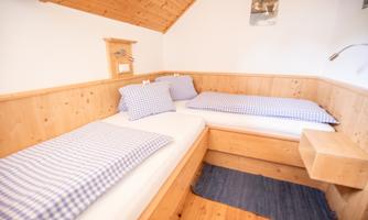 A cozy bedroom with two single beds and wooden paneling. The bedding features a blue and white check pattern and the floor is covered with a soft carpet.