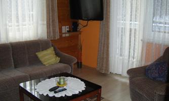 A cozy living room with a sofa, an armchair, and a table. The room has large windows with curtains and a television on the wall.