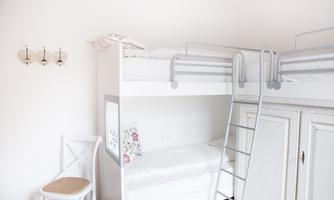 A bright room with a bunk bed and a white wall. A chair and a wardrobe complement the simple furnishings.