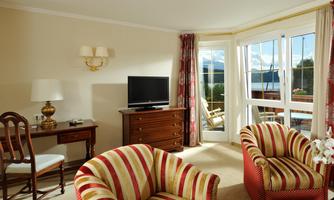 A cozy living room with striped armchairs and a television. Large windows provide a view outside and plenty of daylight.