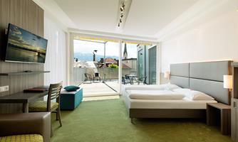 A modern hotel room with a double bed and a large window that leads to a balcony overlooking the city. The room is bright and inviting, with a desk and comfortable chairs.