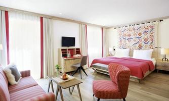 A modern hotel room with a large bed and a cozy seating area. The decor is bright and friendly, with decorative elements on the walls.