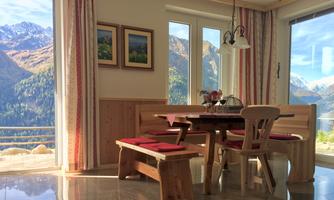 A cozy dining area with wooden furniture and a view of the mountains. Large windows let in plenty of daylight.