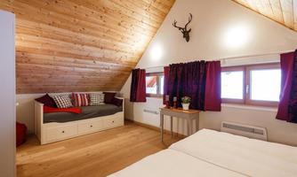 A cozy bedroom with a wooden ceiling and large windows. The furnishings include a tranquil bed and a seating area with colorful cushions.