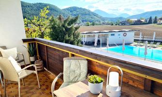 A cozy balcony with a view of a pool and the surrounding mountains. There are plants and two seating areas that invite you to relax.