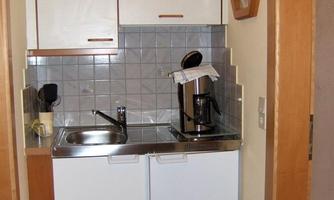 A small kitchenette with a sink, a refrigerator, and a coffee machine. The walls are painted in light colors and there is a trash can in the corner.