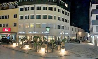 This is Hotel Hecher in 2023. The facade is illuminated with modern lighting, and the outdoor seating area with palm trees creates a welcoming atmosphere.