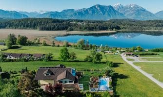 A picturesque landscape with a house, a pool, and a clear lake. In the background, majestic mountains and green meadows can be seen.