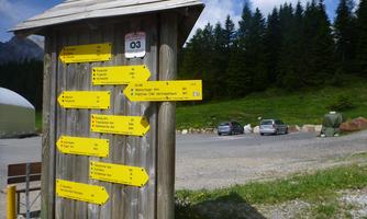 A wooden signpost with yellow signs indicating different directions. In the background, trees and a parking lot can be seen.