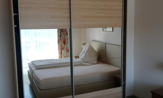 A bright bedroom with a large mirrored wardrobe and two beds. The windows let in a lot of daylight and there are colored curtains.
