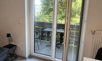 A bright room with an open balcony door that leads to a balcony. Outside, trees and a table with chairs are visible.