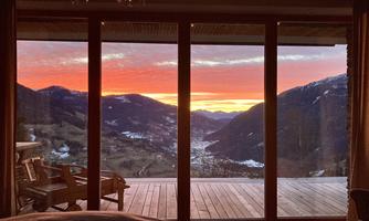 A beautiful sunset over a valley with mountains in the background. The view is enhanced by large glass doors of a chalet.