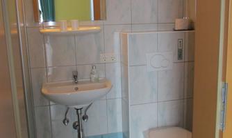 A small bathroom with a shower, a sink, and a toilet. The walls are tiled in light tiles, and there is a mirror above the sink.
