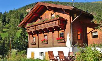 A traditional wooden house with flower boxes and a sunny balcony. In the foreground, there are wooden chairs on a small terrace, surrounded by beautiful nature.