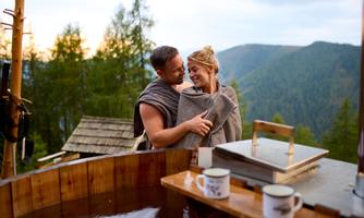 A couple embracing in a warm wooden bath in nature. In the background, there are mountains and trees, and the atmosphere is romantic and relaxing.