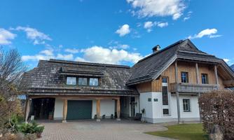 A modern house with a spacious garage area and a distinctive roof structure. The sky is blue with a few clouds, and the surroundings are well-maintained.