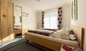 A bright bedroom with a double bed and floral patterns on the bedding. The walls are in neutral colors and large windows provide plenty of daylight.