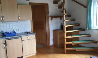 A cozy room with a small kitchen and a wooden staircase. The walls are light, and large windows let in a lot of light.