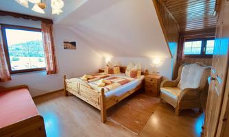 A cozy bedroom with a double bed, an armchair, and a view through large windows. The room is decorated in warm wood tones and has a friendly atmosphere.