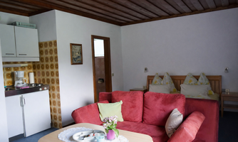 A cozy living space with a red couch and a table. In the background, a small kitchenette and a double bed are visible.