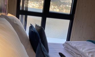 A cozy bed with pillows and a view of a lake and the mountains through a large window. The view is clear and shows a peaceful, natural landscape.