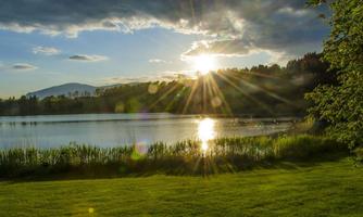 A tranquil lake at sunset, surrounded by green meadows and trees. The sun shines over the water, creating a warm play of light.