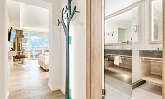A bright, modern room with a large window and access to a balcony. In the foreground, an elegant coat rack and a stylish bathroom are visible.
