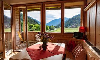 A cozy room with large windows that offer a view of green mountains and valleys. The table is decorated with a red cloth and a flower vase.